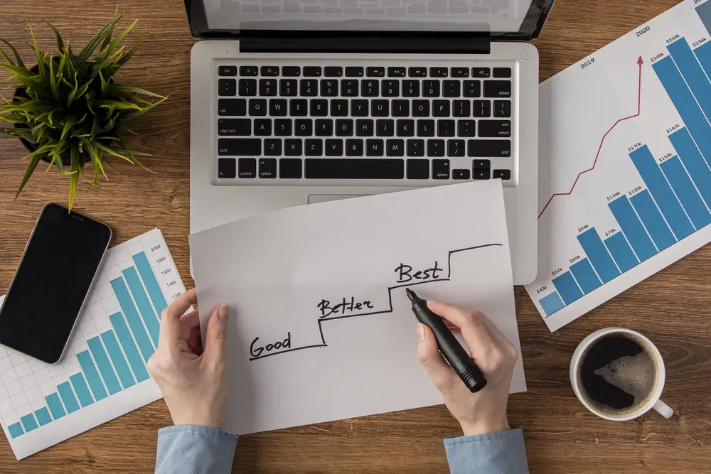 A person writing the words "Good, Better, Best" on a piece of paper on a desk with a laptop, phone and pieces of paper next to it.