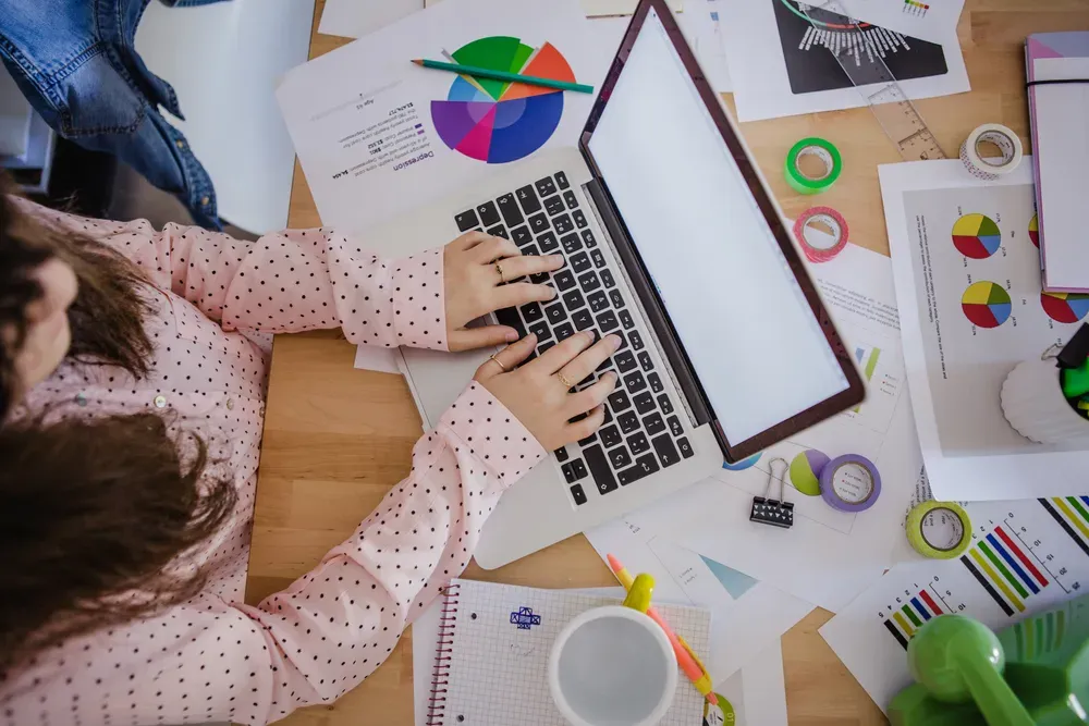 A woman working on a laptop and the desk is full of sheets of paper under the laptop.